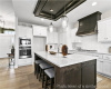 Kitchen featuring appliances with stainless steel finishes, custom exhaust hood, recessed lighting, a raised ceiling, and a kitchen island with sink